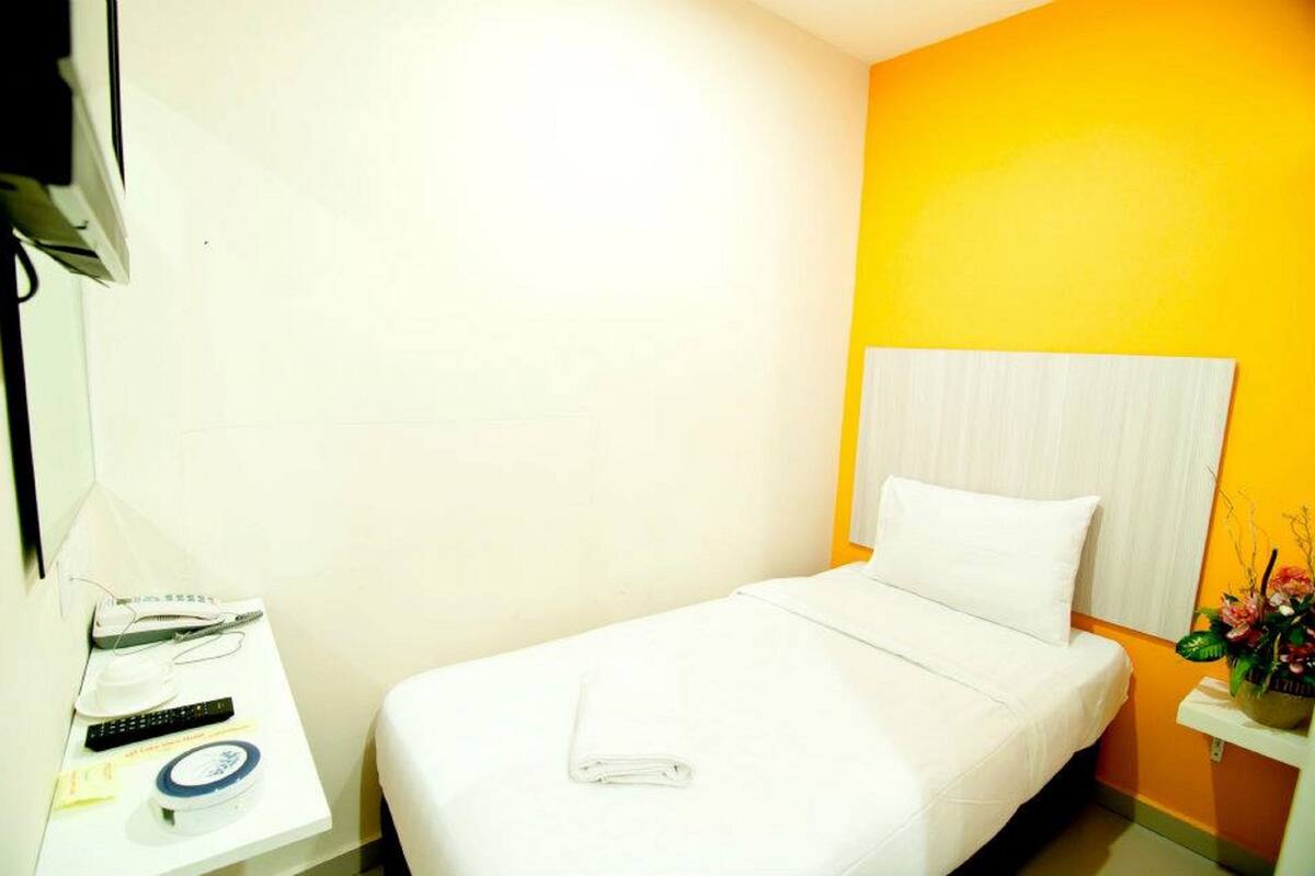 A hotel room with a single bed, a bedside table with a remote control, a clock, and a small plant.