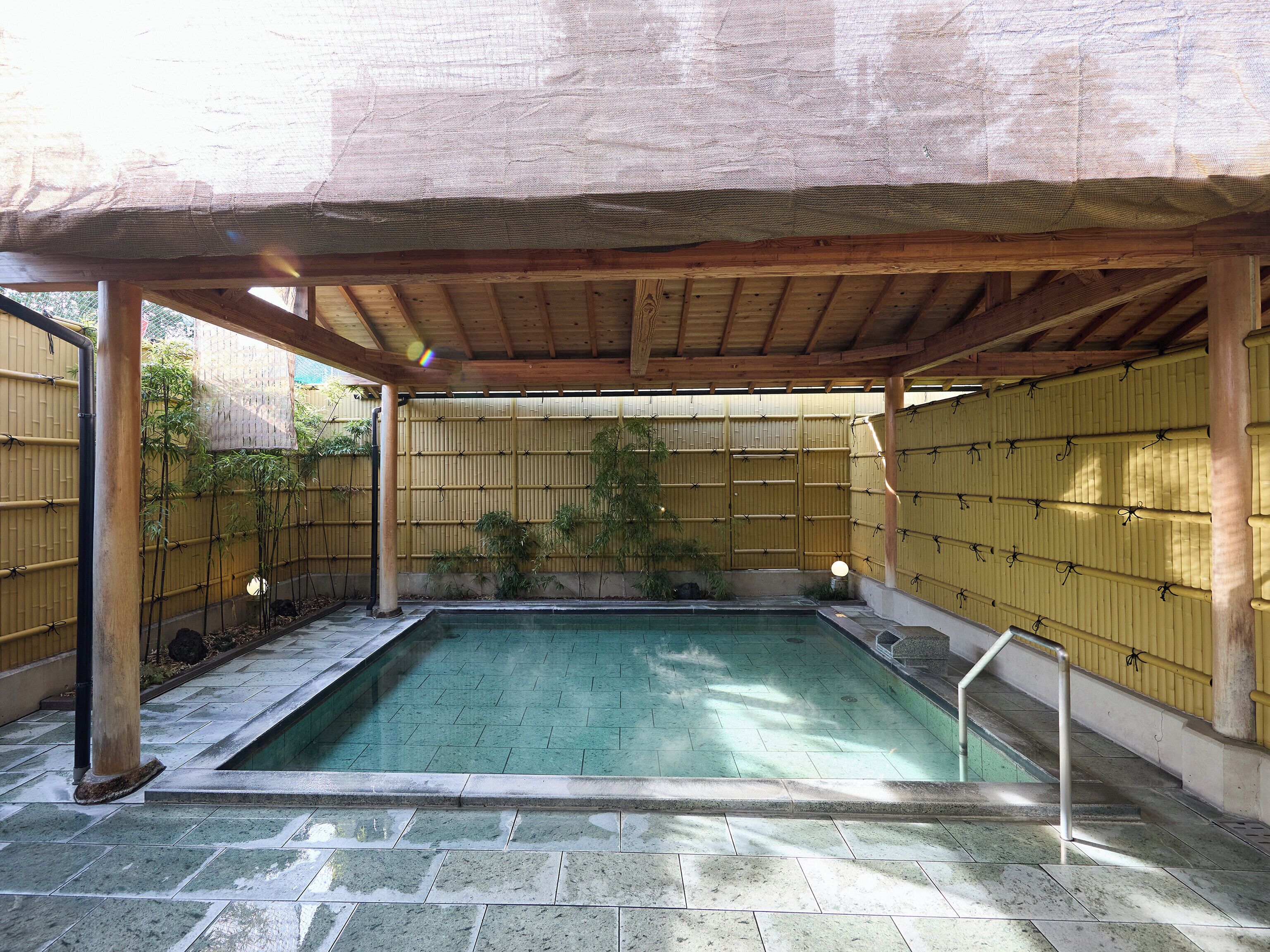 A covered outdoor swimming pool with a bamboo fence, wooden beams, and tiled flooring.