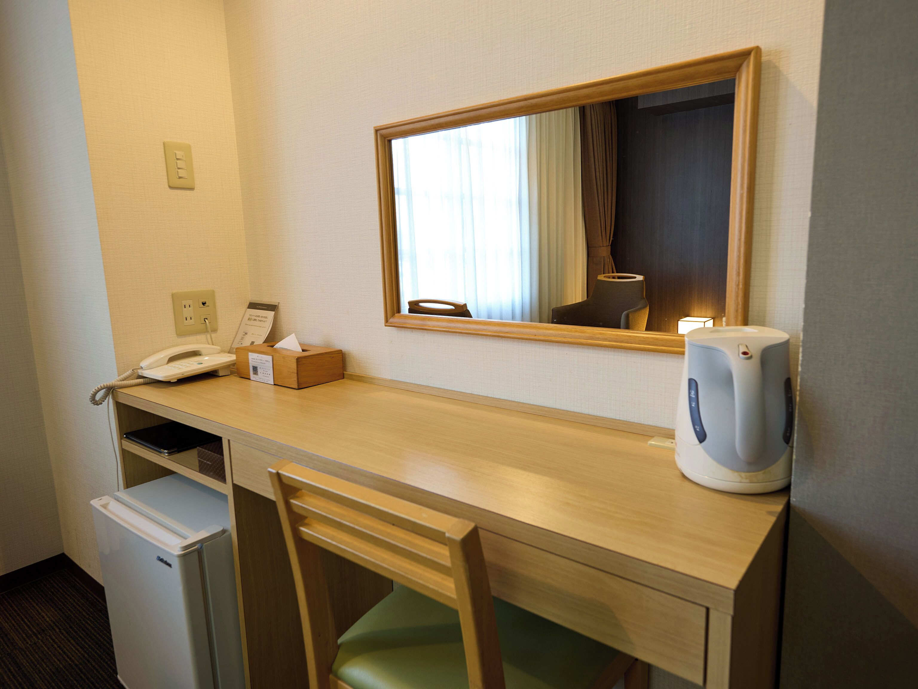A hotel room with a wooden desk, a chair, a telephone, a tissue box, a kettle, and a mirror.