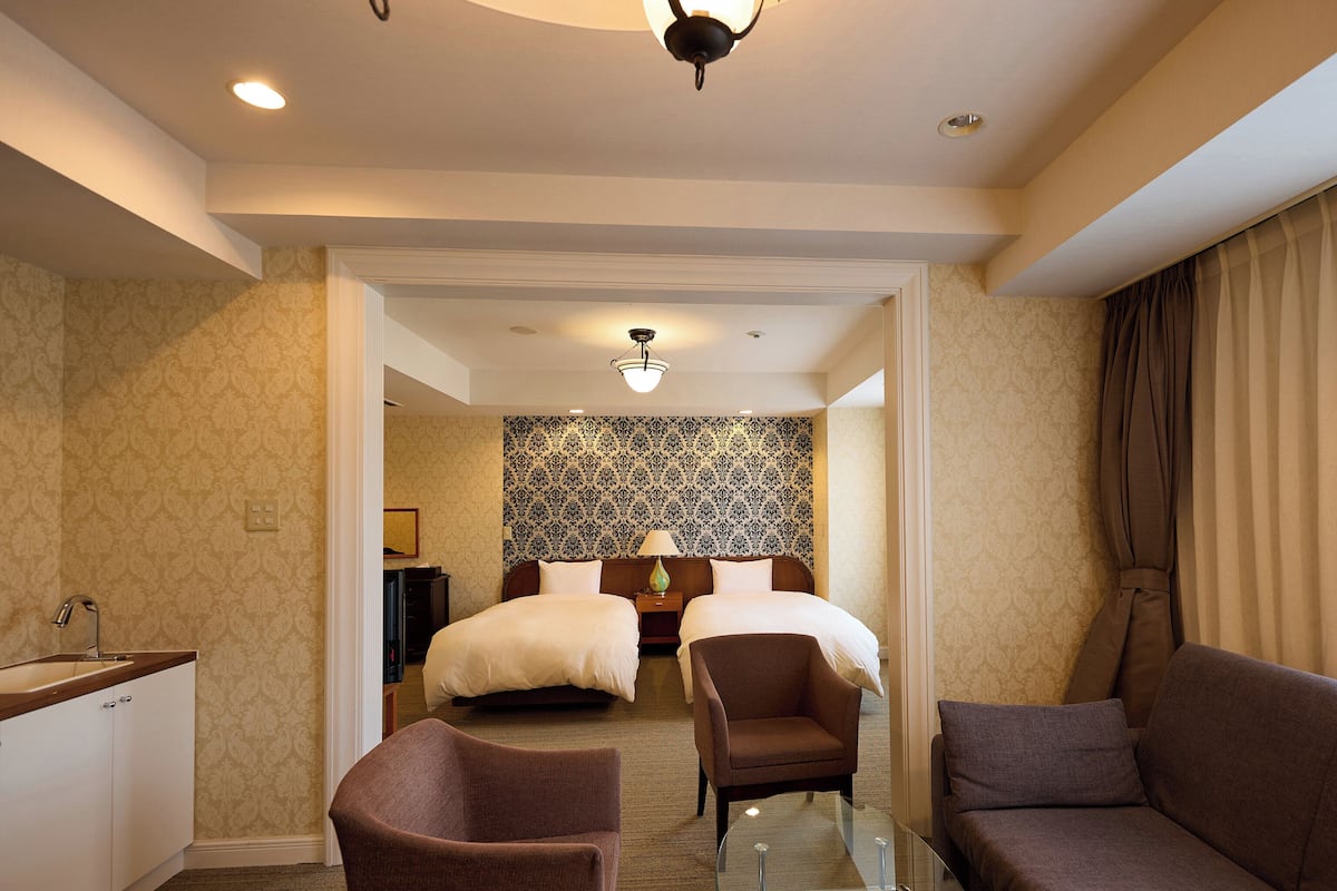 A hotel room with a bed, two armchairs, a glass coffee table, a desk with a sink, and a patterned wallpaper.