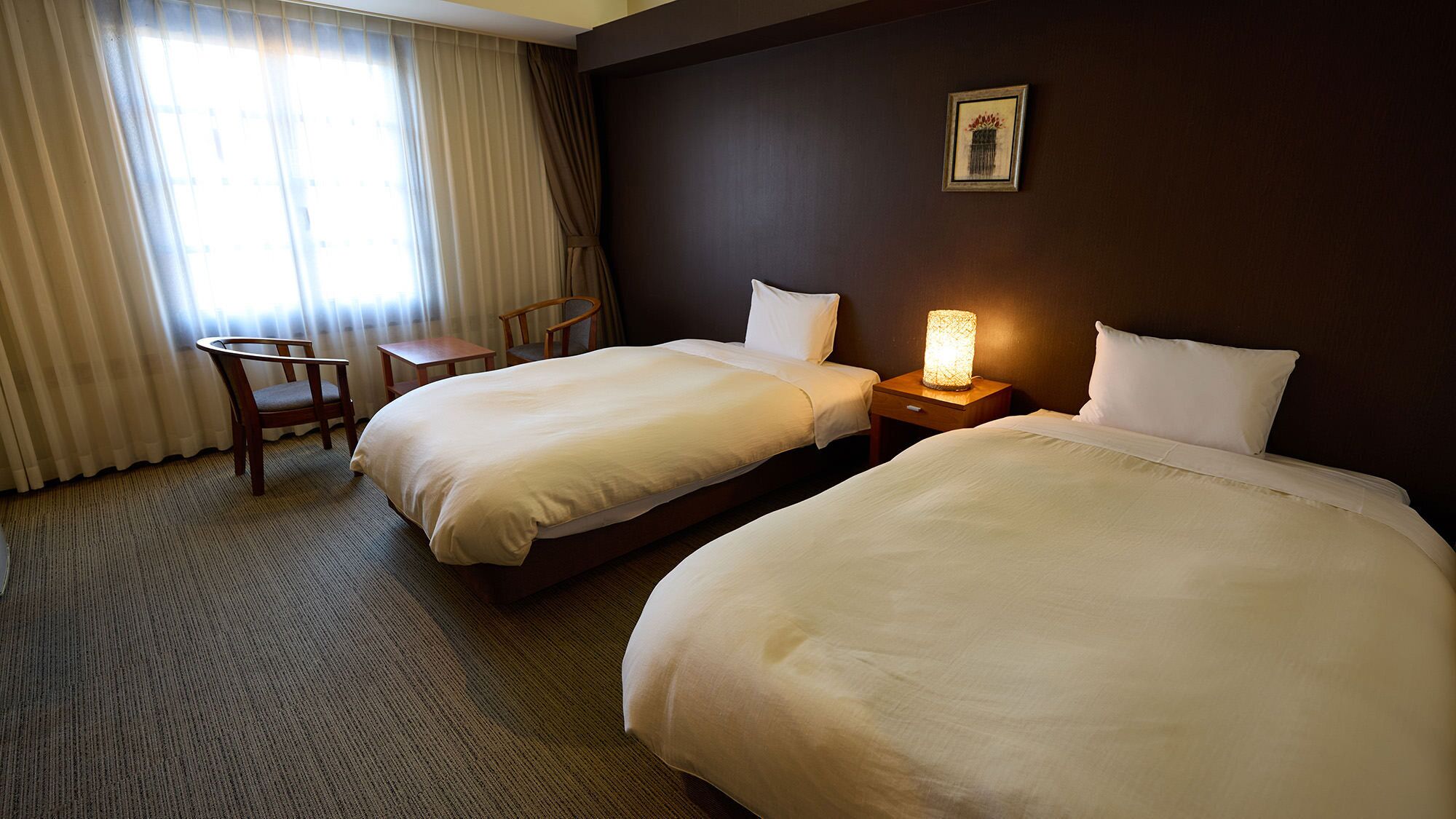 A hotel room with two beds, a desk, a chair, and a nightstand with a lamp.