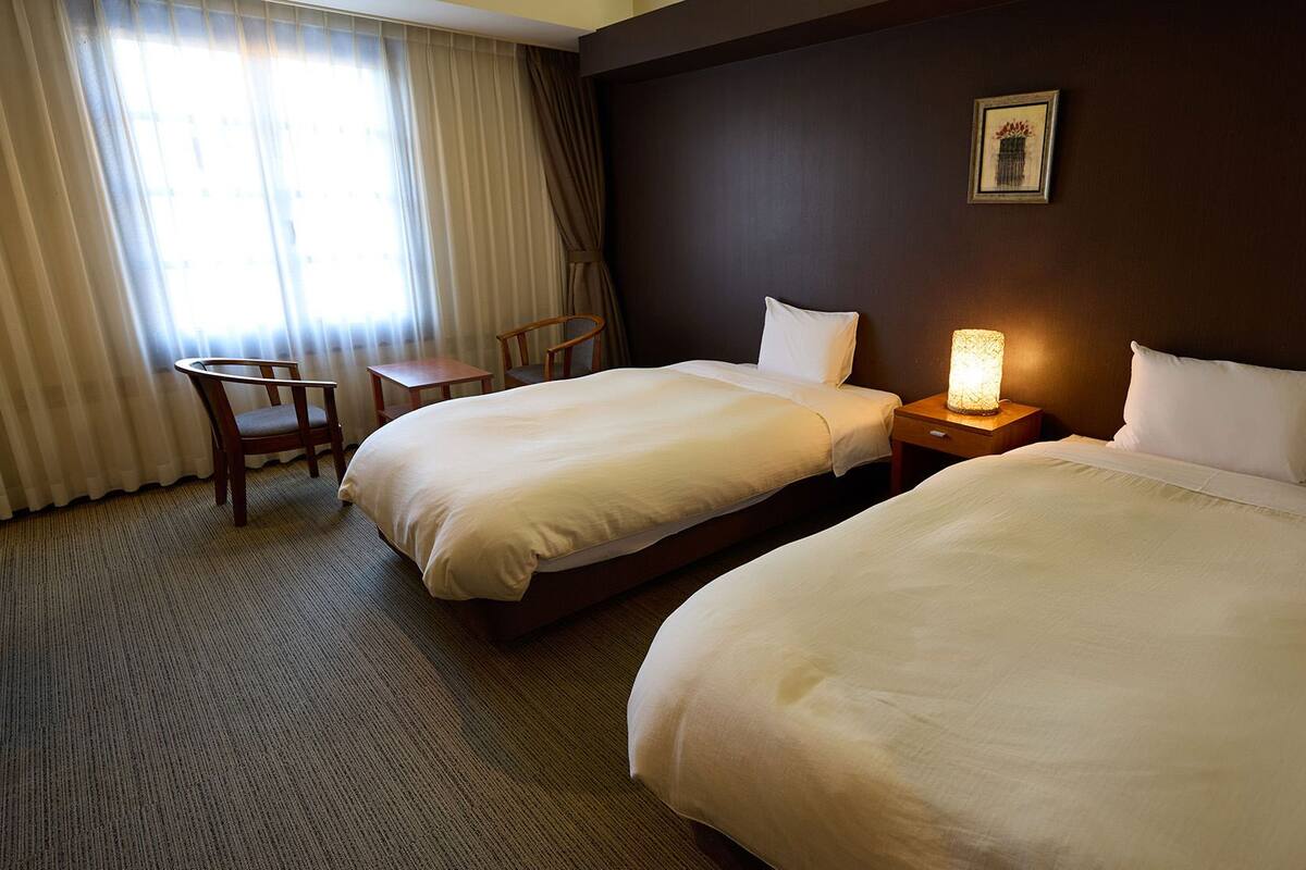 A hotel room with two beds, a desk, a chair, and a nightstand with a lamp.