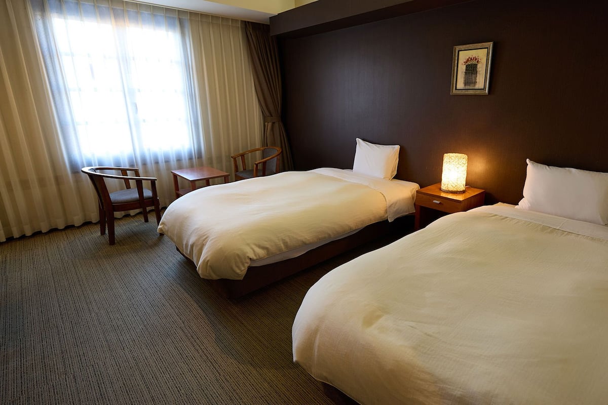 A hotel room with two beds, a desk, a chair, and a nightstand with a lamp.