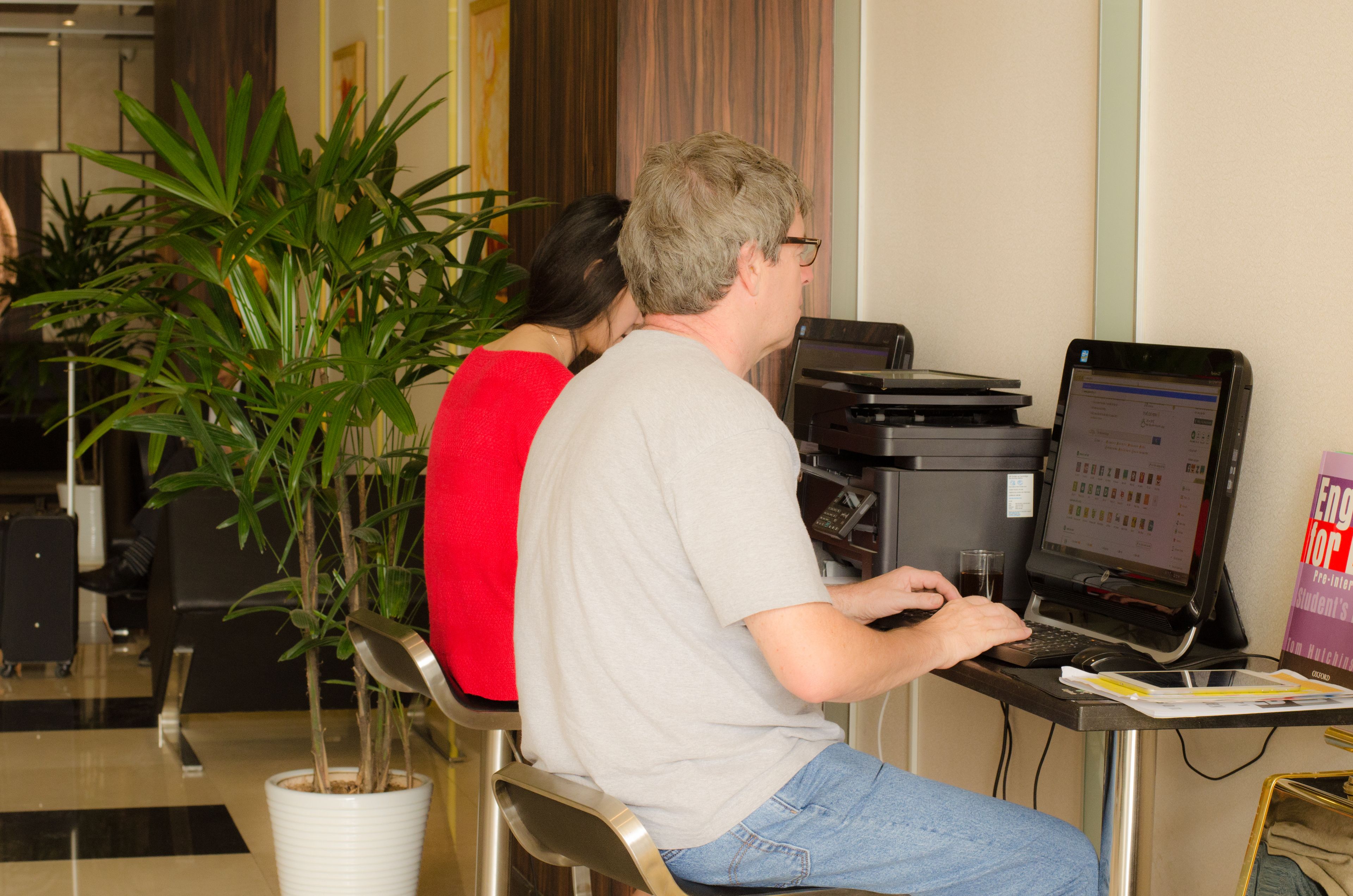 A man using a laptop at a desk with a computer monitor, a potted plant, and a book on the table.