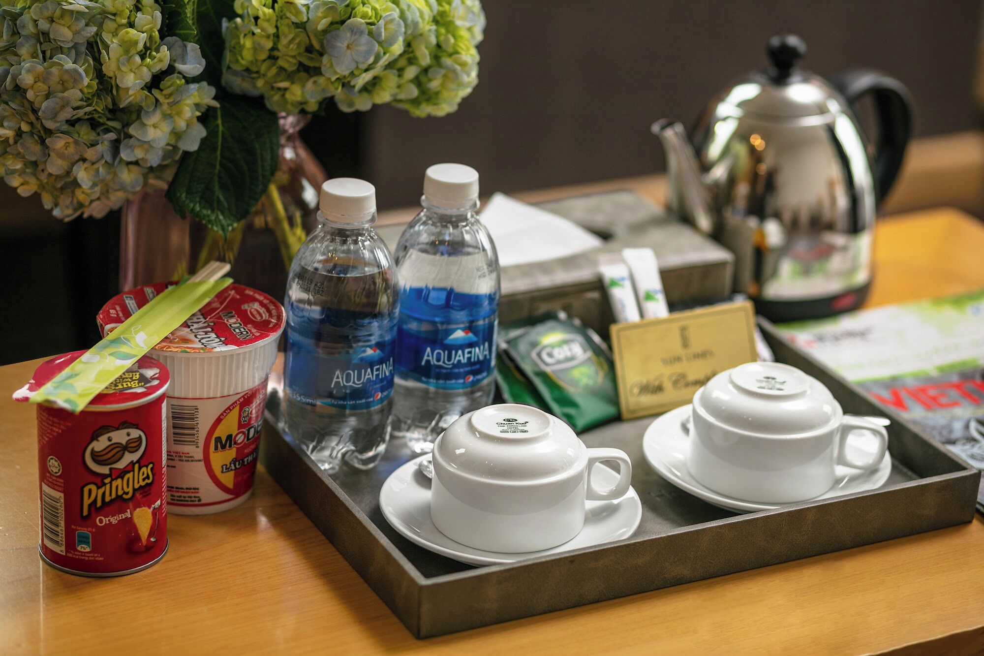 A tray with a can of Pringles, two bottles of Aquafina water, a coffee pot, and two white cups on a wooden table.