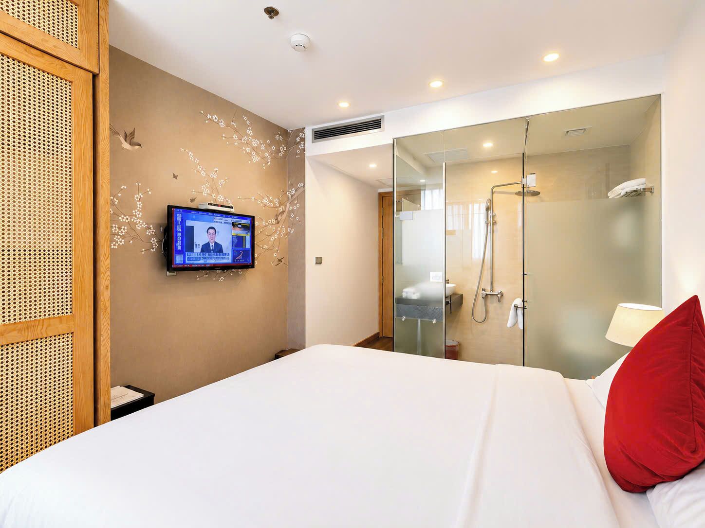 Habitación de hotel con una cama, un televisor fijado a la pared, ducha con cerramiento de vidrio y un separador de madera con diseño perforado.