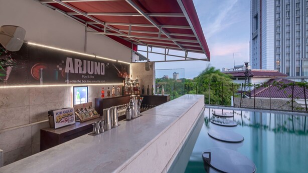 Zona de bar junto a la piscina con un letrero que dice "ARJUNO Pool & Bar", diversas botellas y vasos, y vista de un paisaje urbano con edificios y árboles.