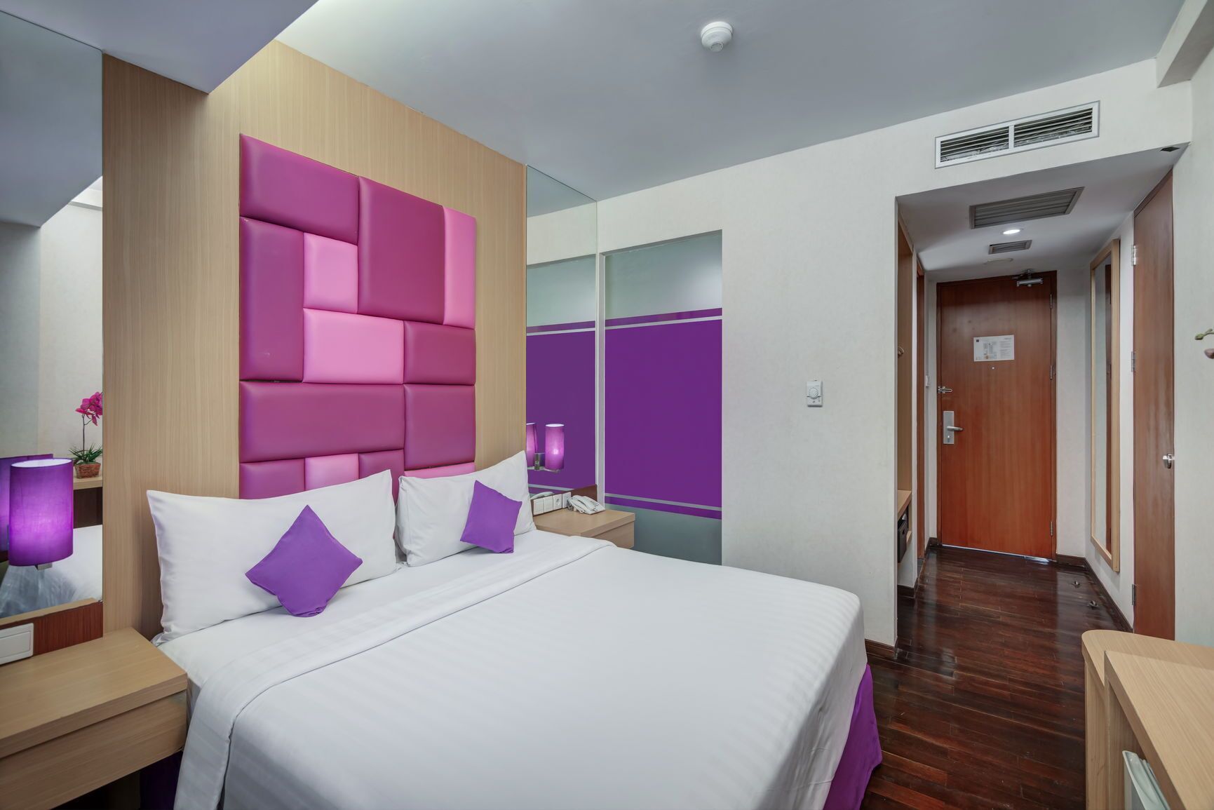Habitación de hotel con una cama grande, cabecera de madera, mesita de noche con lámpara y baño visible a través de un mamparo de vidrio.