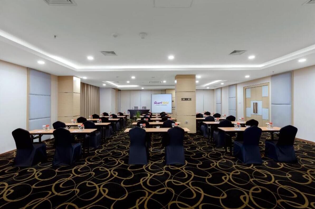 Un salón de conferencias con una pantalla grande al frente, varias mesas con sillas y piso alfombrado.