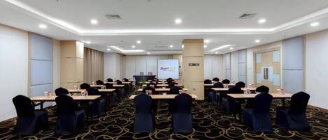 Un salón de conferencias con una pantalla grande al frente, varias mesas con sillas y piso alfombrado.