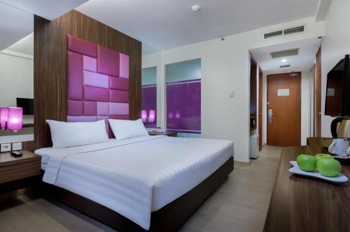 A modern hotel room with a large bed, a wooden headboard, a bedside table with a lamp, and a dining area with a table and chairs.
