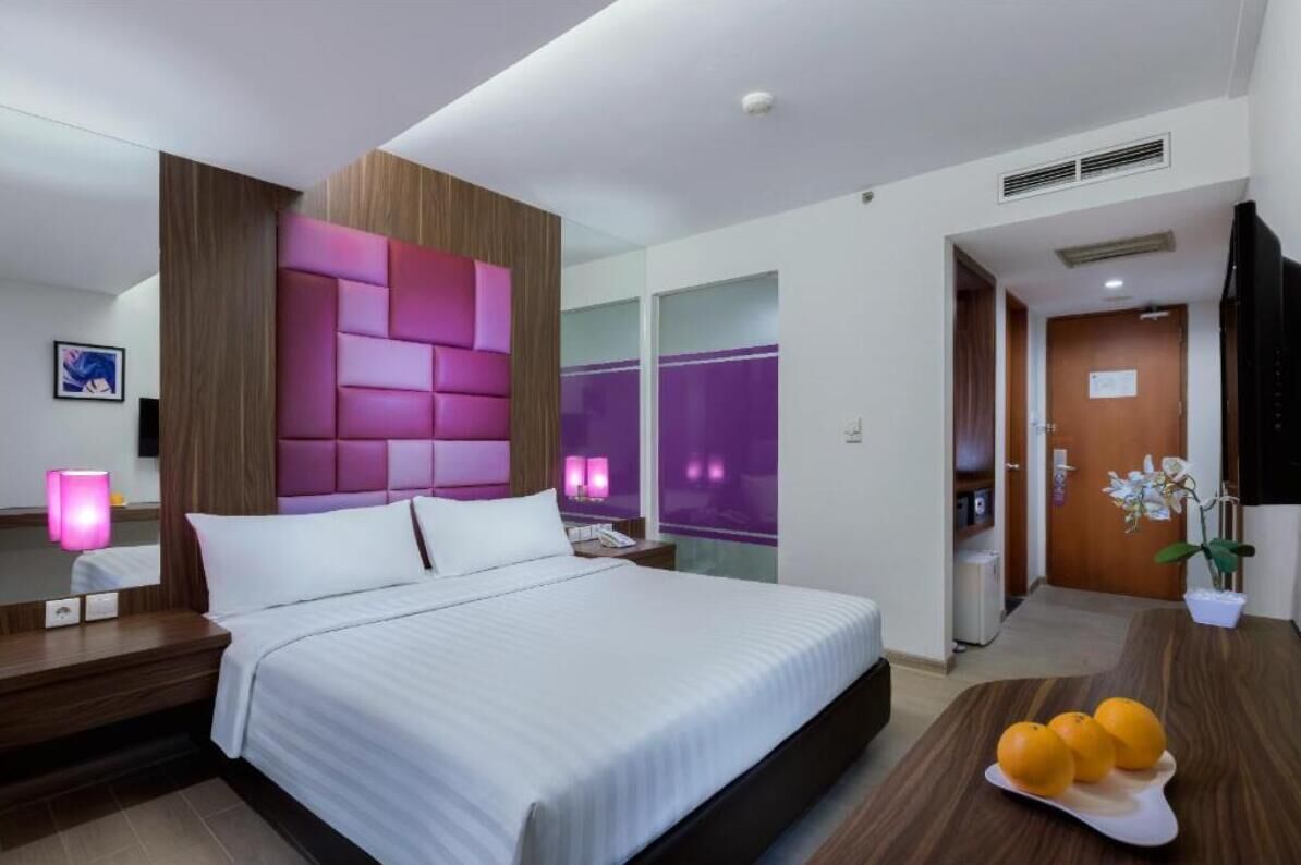 Une chambre d’hôtel moderne avec un grand lit, une tête de lit en bois, une table de chevet avec une lampe et une petite table sur laquelle sont posées des oranges.