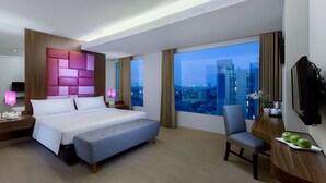 Una habitación de hotel moderna con una cama grande, una zona de estar, un escritorio con silla y un ventanal con vistas a la ciudad.