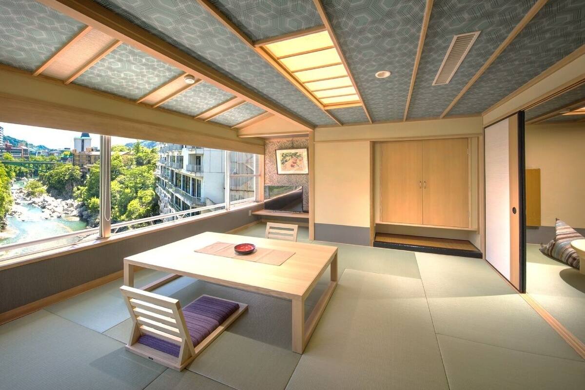 A modern interior with a large window, a wooden table, tatami flooring, and a view of a natural landscape.
