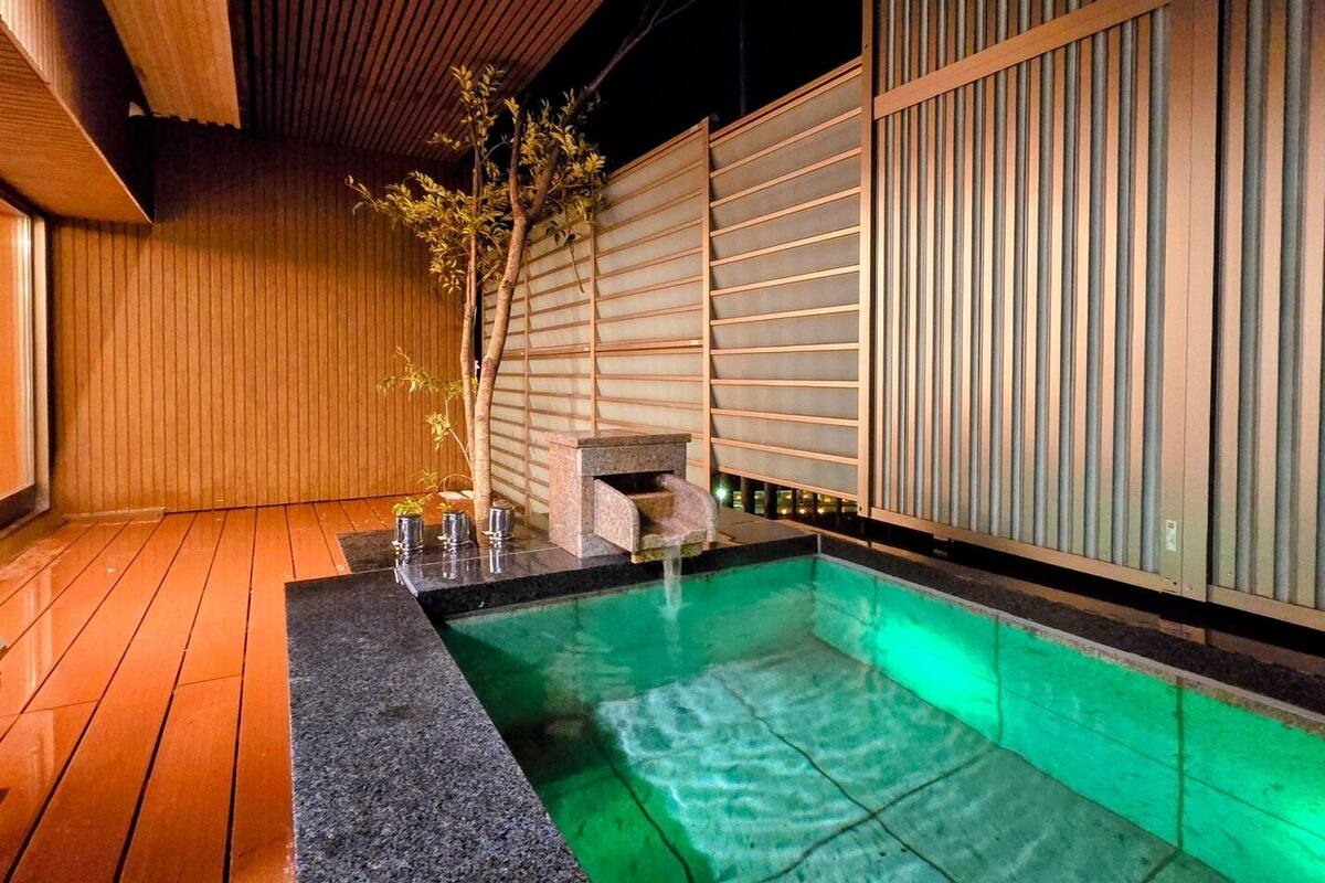 A traditional Japanese-style outdoor hot spring with wooden flooring, a stone bench, and a wooden fence.
