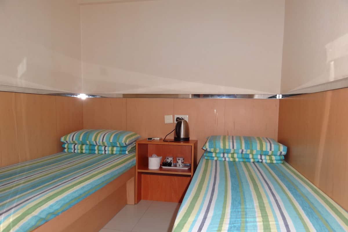 A small, compact room with two single beds, each with striped bedding. There is a wooden shelf unit in the center with a kettle and a small table.