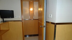 Quarto de hotel com porta de madeira, televisão sobre um suporte e interruptor de luz na parede.