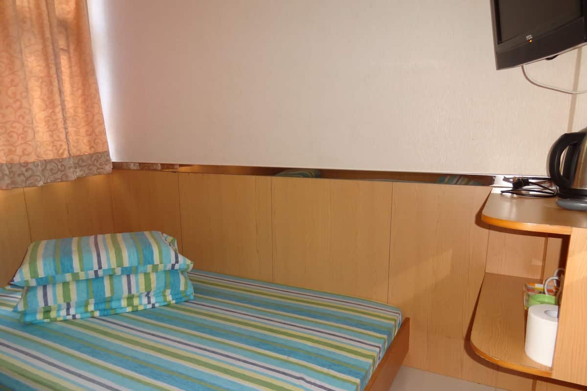 A small, single-bed room with a striped bedspread, a wooden headboard, a television mounted on the wall, and a small shelf with toiletries.