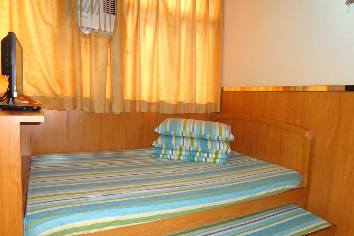 A single bed with a striped mattress, a wooden headboard, a bedside table with a lamp, and a window with curtains.