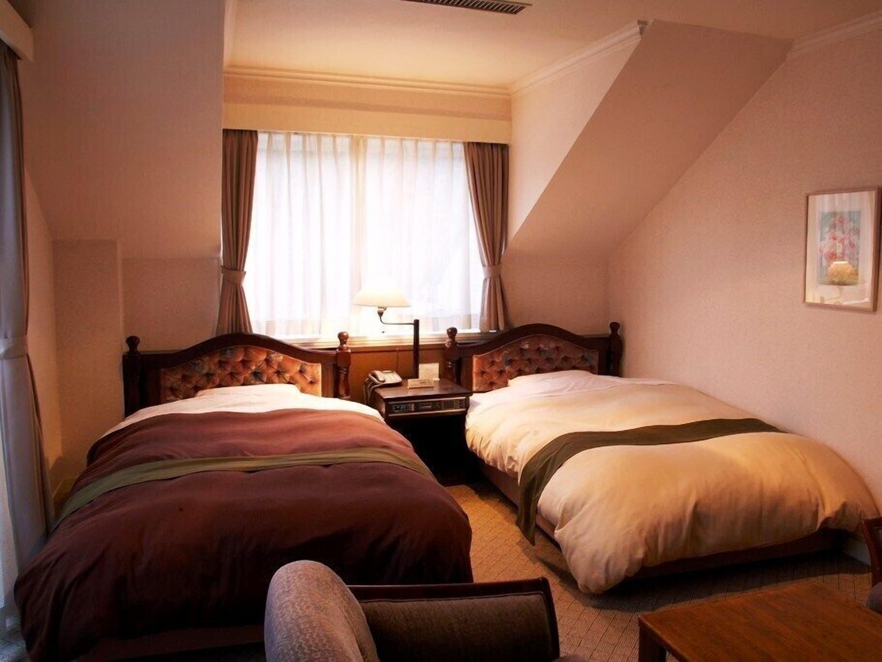 A hotel room with two beds, a nightstand, a lamp, and a window with curtains.