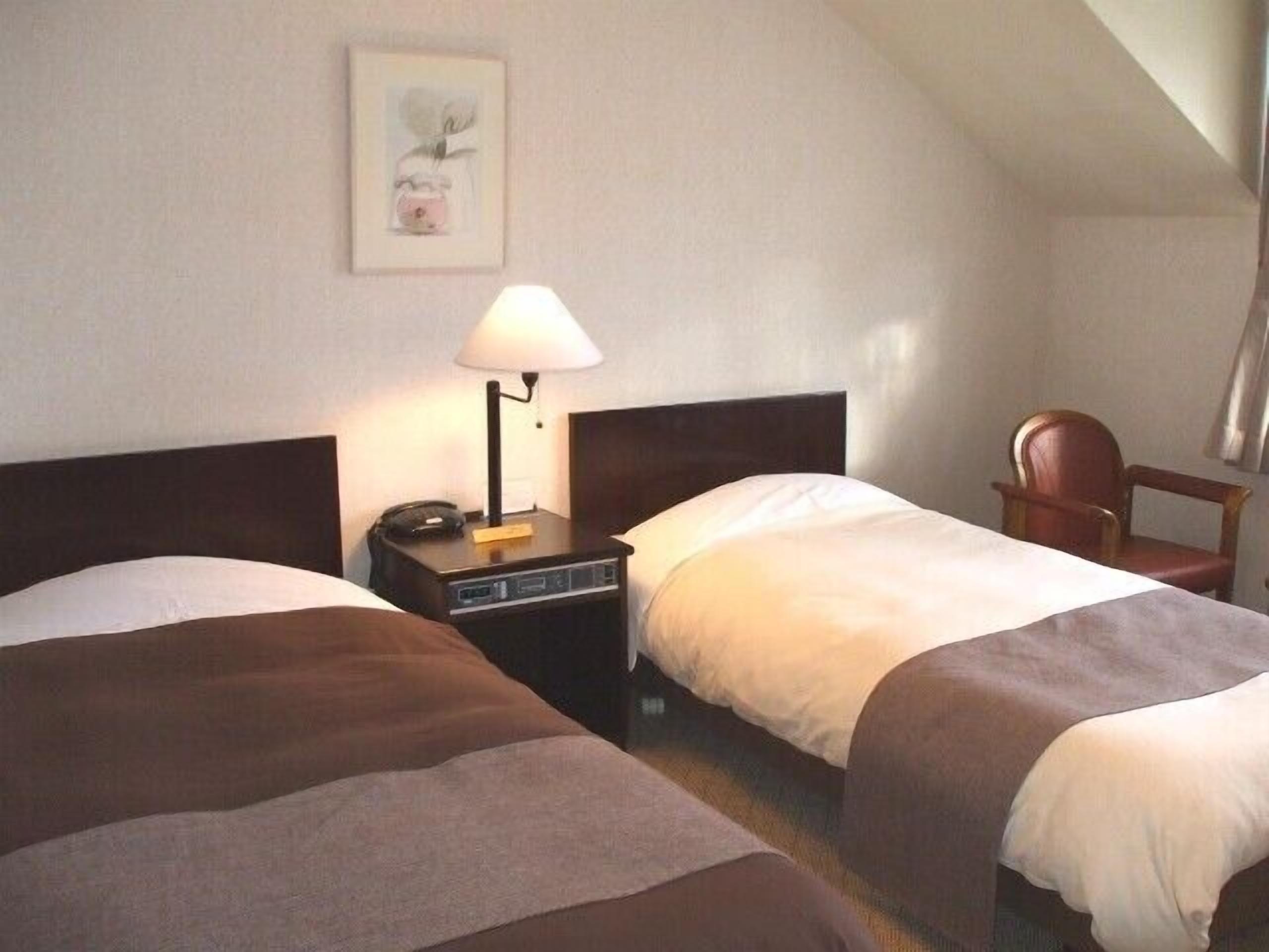 Habitación de hotel con dos camas, una mesita de noche con lámpara, una silla e imagen enmarcada en la pared.