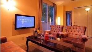 Habitación de hotel con televisor de pantalla plana montado en la pared, un ventanal con cortinas, un sillón cómodo, un sofá y una pequeña mesa con una botella y flores.