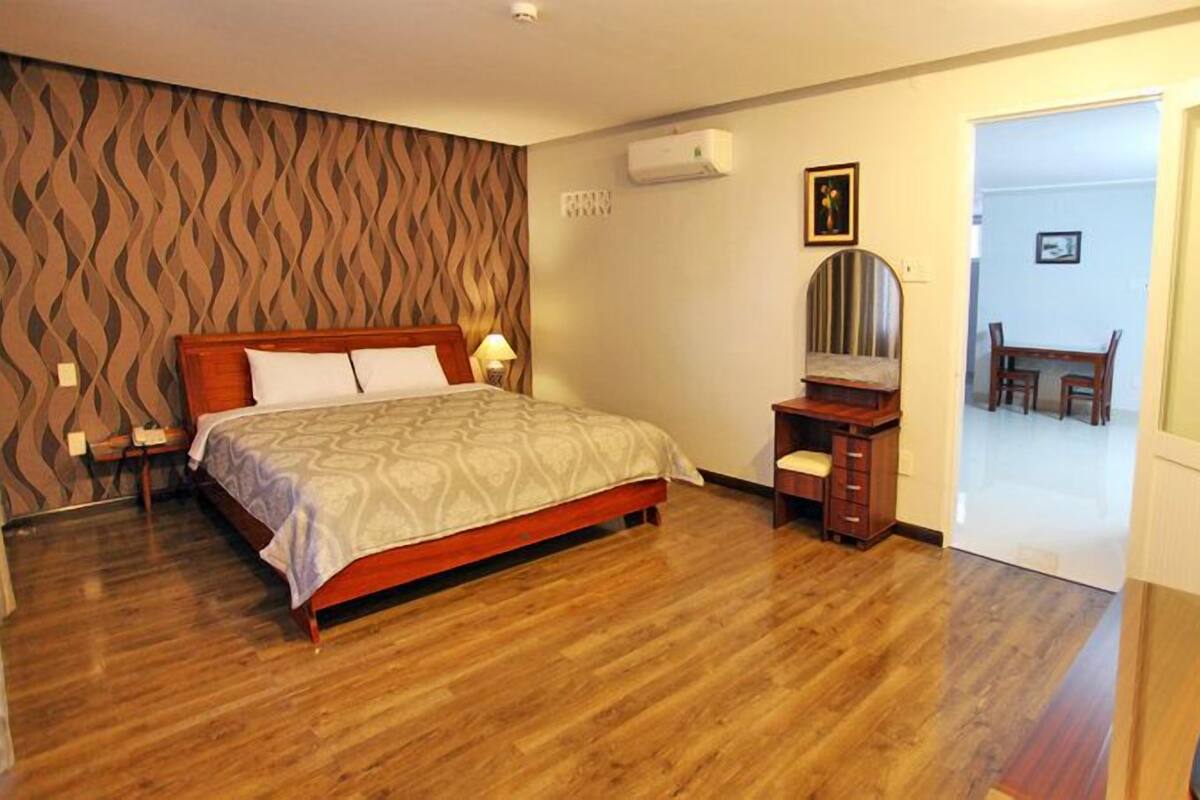 A bedroom with a wooden bed, a desk with a mirror, and a door leading to a dining area.