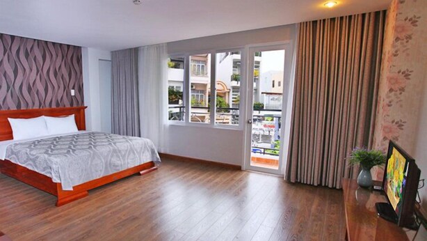 A hotel room with a wooden bed, a balcony with curtains, and a TV on a wooden table. - White Swan Apartment (Nha Trang)