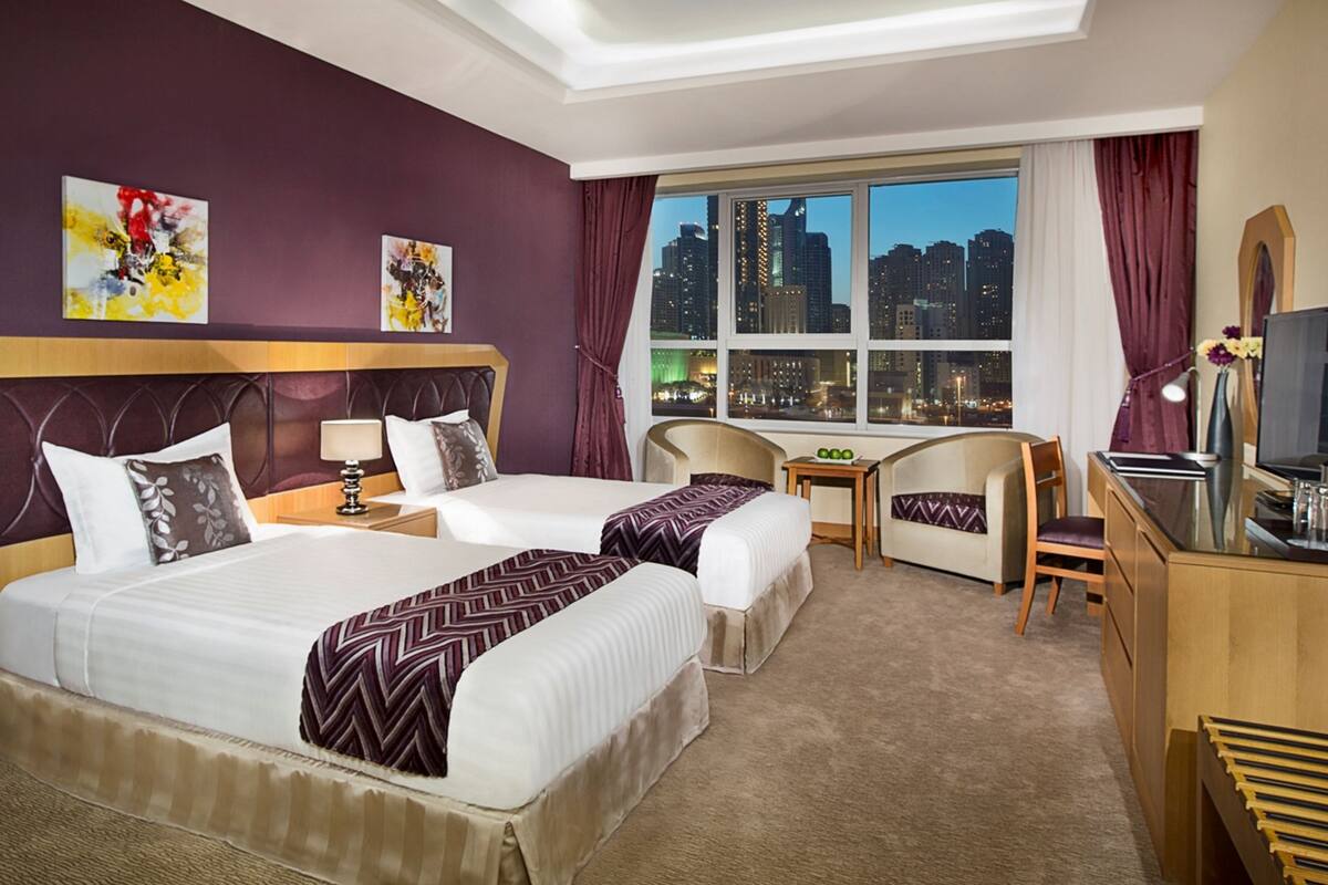 A hotel room with two beds, a desk, a chair, a TV, and a window with a city view.