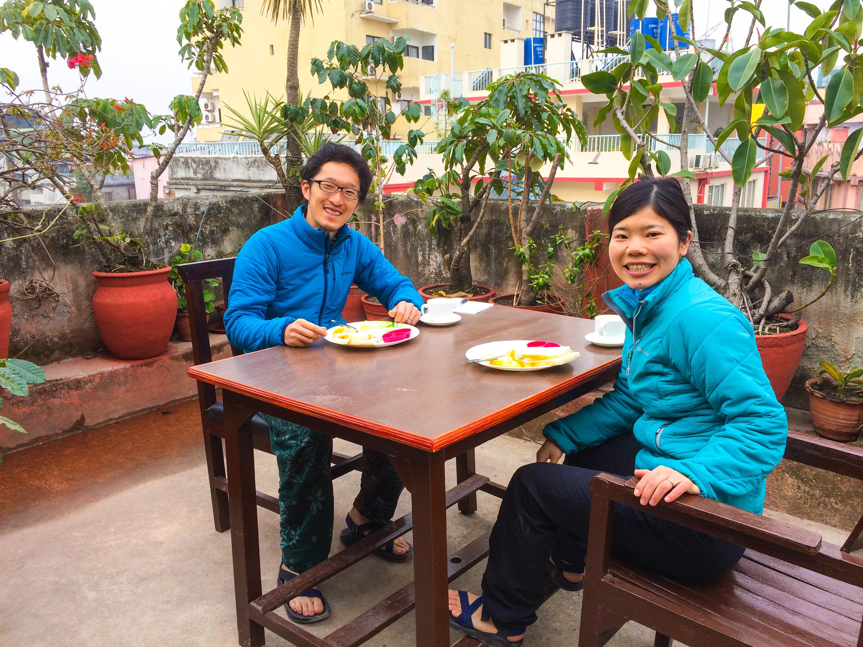 Two people sitting at a wooden table outdoors with potted plants around them.