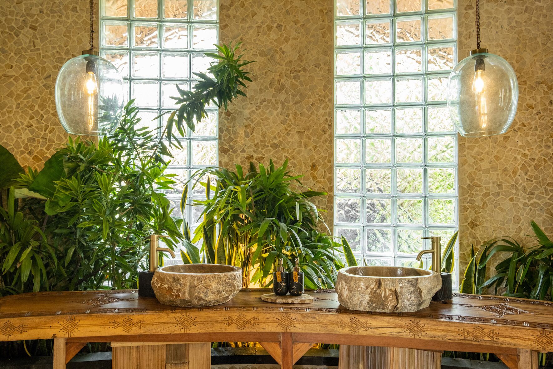 Une table en bois, sur laquelle reposent deux bols en pierre, est entourée d’une végétation luxuriante. Deux globes de verre suspendus, contenant des ampoules, complètent l’ensemble.