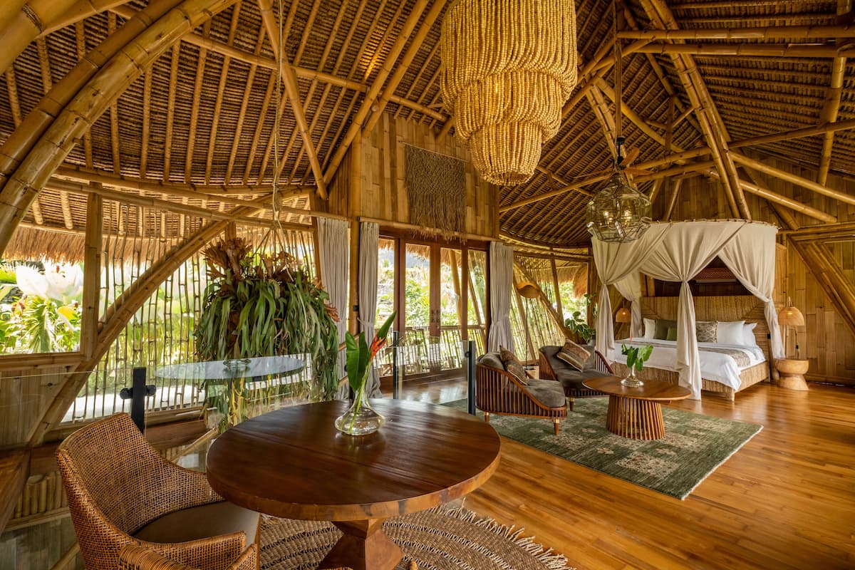 A spacious room with a high ceiling, a large round wooden table, wicker furniture, a canopy bed, and a view of greenery outside.