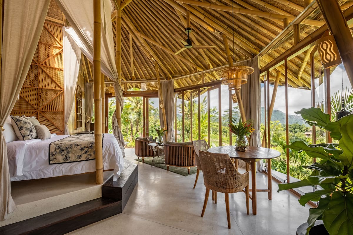A spacious room with a bed, a dining table, and chairs, large windows offering a view of the outdoors.