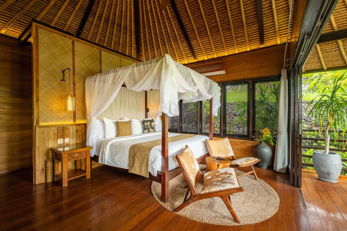 A spacious bedroom with a canopy bed, wooden furniture, and a large window offering a view of greenery.