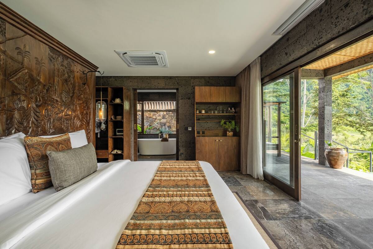 A bedroom with a large bed, a wooden headboard, a bathroom with a bathtub, and a balcony with a view of greenery.