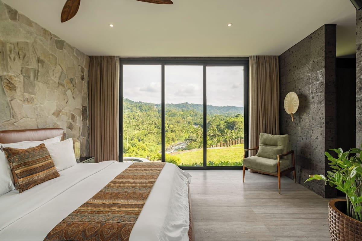 A bedroom with a large bed, a view of a green landscape, a stone wall, and a modern chair.
