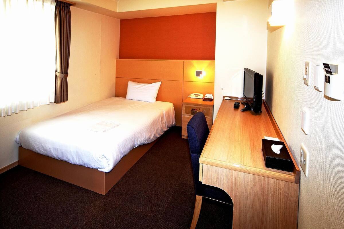 A hotel room with a bed, a desk with a computer, a telephone, and a wall-mounted TV.
