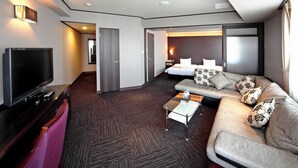 Quarto de hotel moderno com televisão de tela plana, sofá com almofadas estampadas, uma pequena mesa de centro e cama com roupa de cama branca.