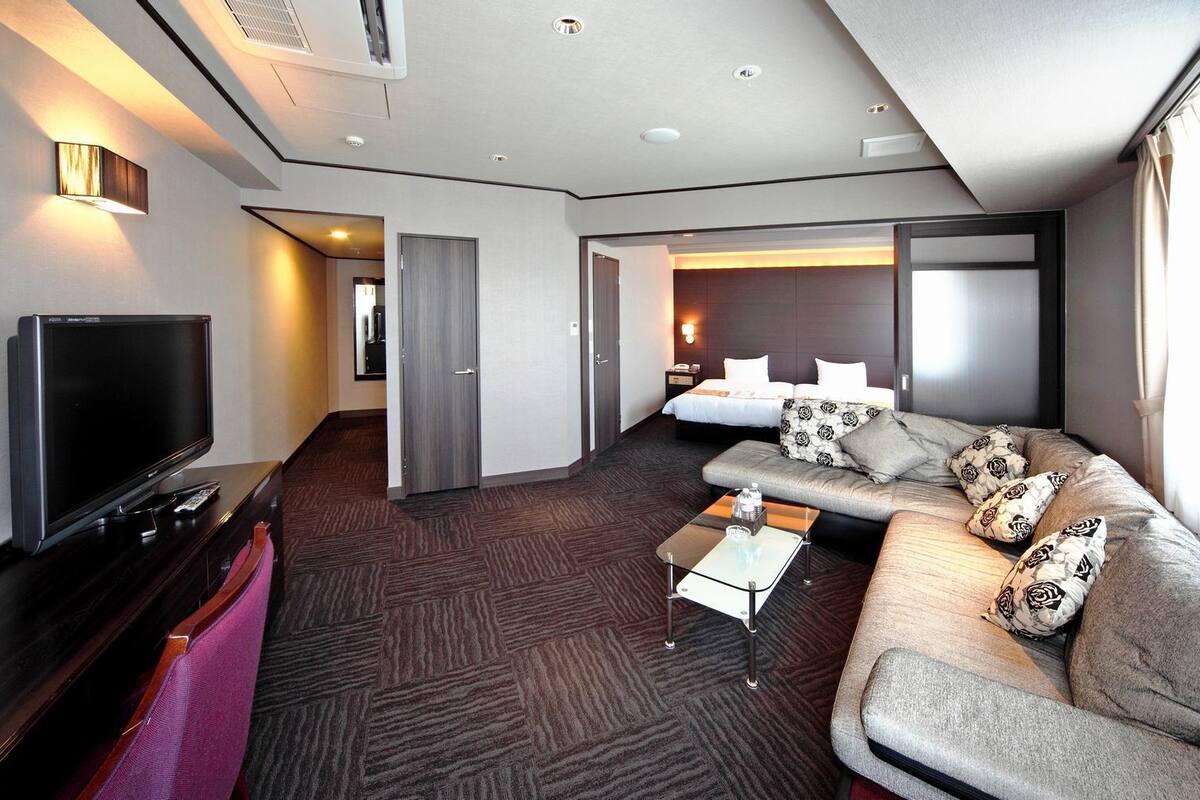 A modern hotel room with a flat-screen TV, a sofa with patterned cushions, a small coffee table, and a bed with white bedding.