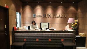 Balcão de recepção de hotel com um recepcionista, uma placa com a inscrição 'Hotel Sun Plaza' e um monitor de computador.
