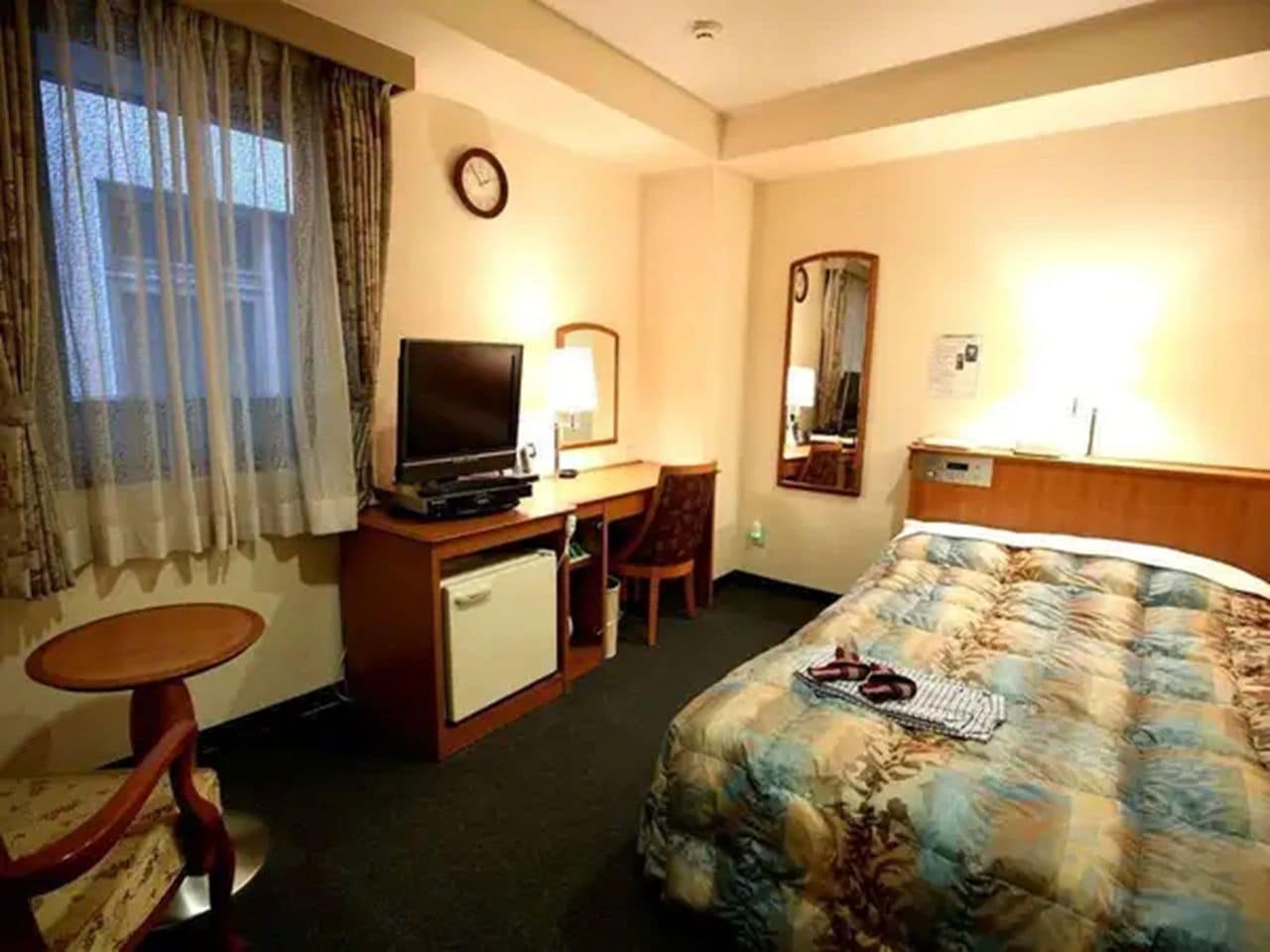 A hotel room with a bed, a desk with a television, a chair, a small table, and a mirror.
