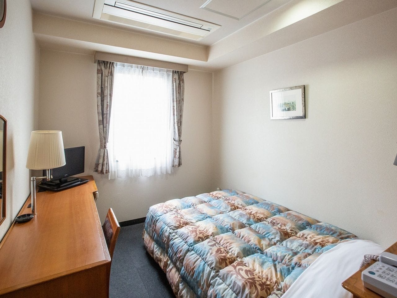 A hotel room with a bed, a desk with a lamp, a television, and a window with curtains.