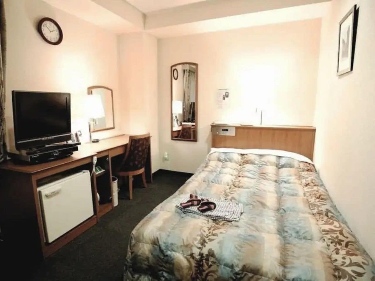 A hotel room with a bed, a desk with a television, a refrigerator, and a mirror.