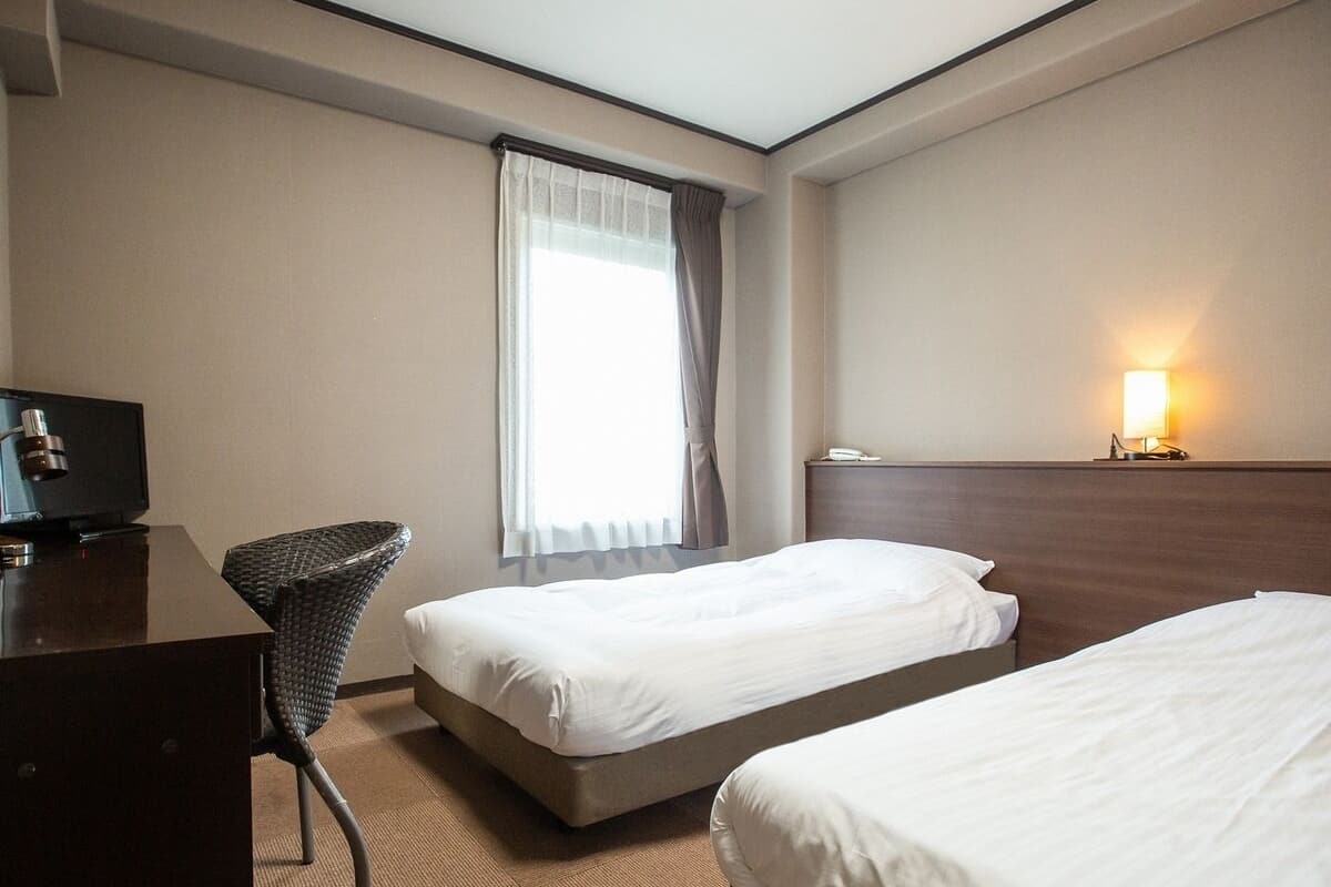 A hotel room with two single beds, a desk with a chair, a television, and a window with curtains.