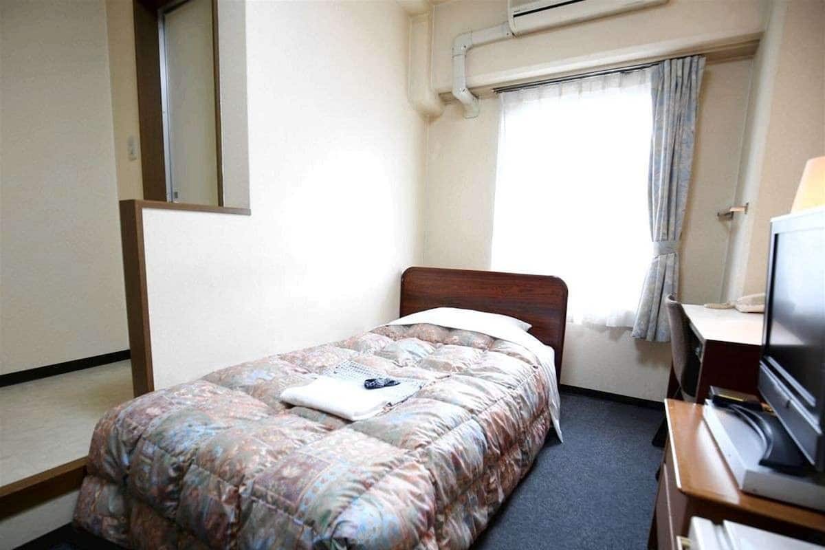 A single bed with a floral comforter, a wooden headboard, a desk with a television, and a window with curtains.