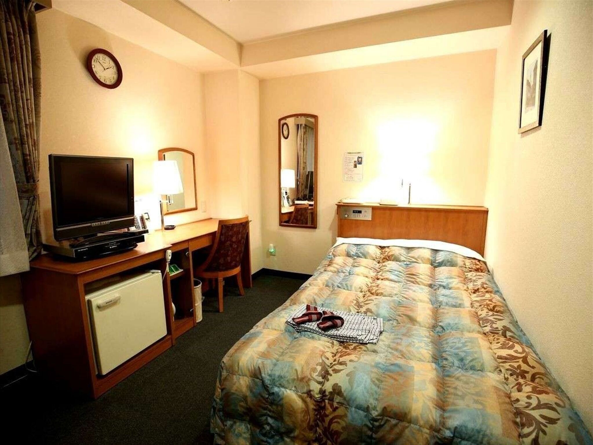 Hotellihuone, jossa on sänky, työpöytä, jossa on televisio, tuoli, pieni jääkaappi, kello ja peili.