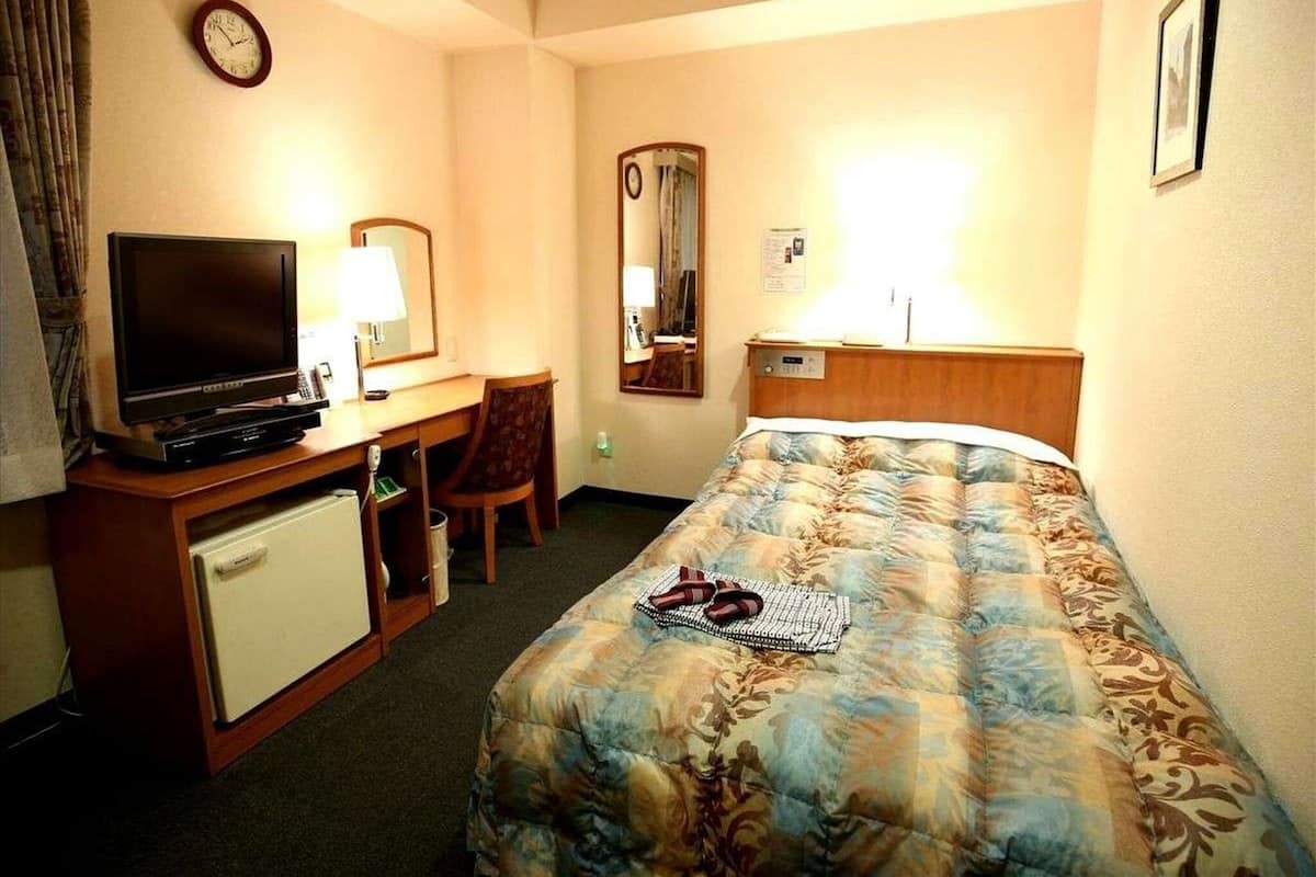 A hotel room with a bed, a desk with a television, a chair, a small refrigerator, and a clock on the wall.