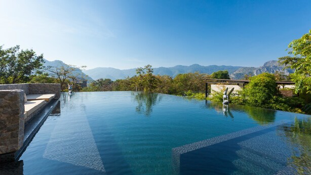 A clear blue infinity pool with a stone bench and a small statue, surrounded by lush greenery and mountains in the background. - Botanica Khao Yai (Pak Chong)