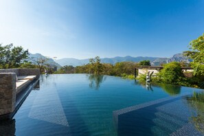 A clear blue infinity pool with a stone bench and a small statue, surrounded by lush greenery and mountains in the background. - Botanica Khao Yai (Pak Chong)