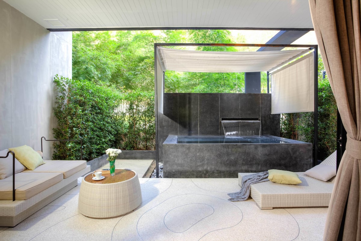 A modern outdoor relaxation area with a hot tub, seating, and a view of lush greenery.